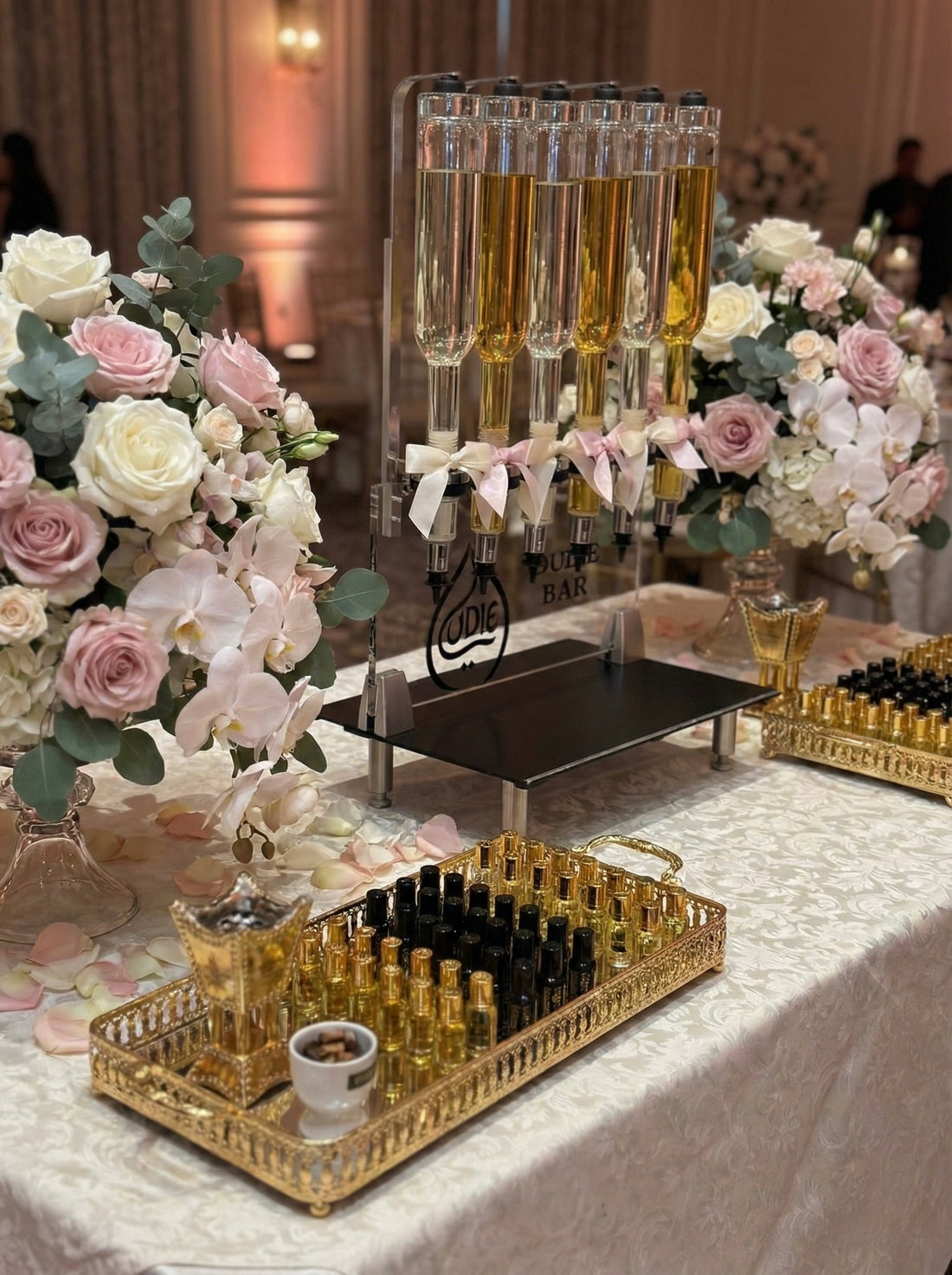 An intimate close-up of a sophisticated "Oud Bar" setup on a table covered with a patterned cloth. The dispenser with amber liquids is flanked by gorgeous bouquets of pink and white roses and orchids. Two ornate gold trays hold an array of black and gold perfume bottles, alongside a small burner, all under soft, warm lighting.