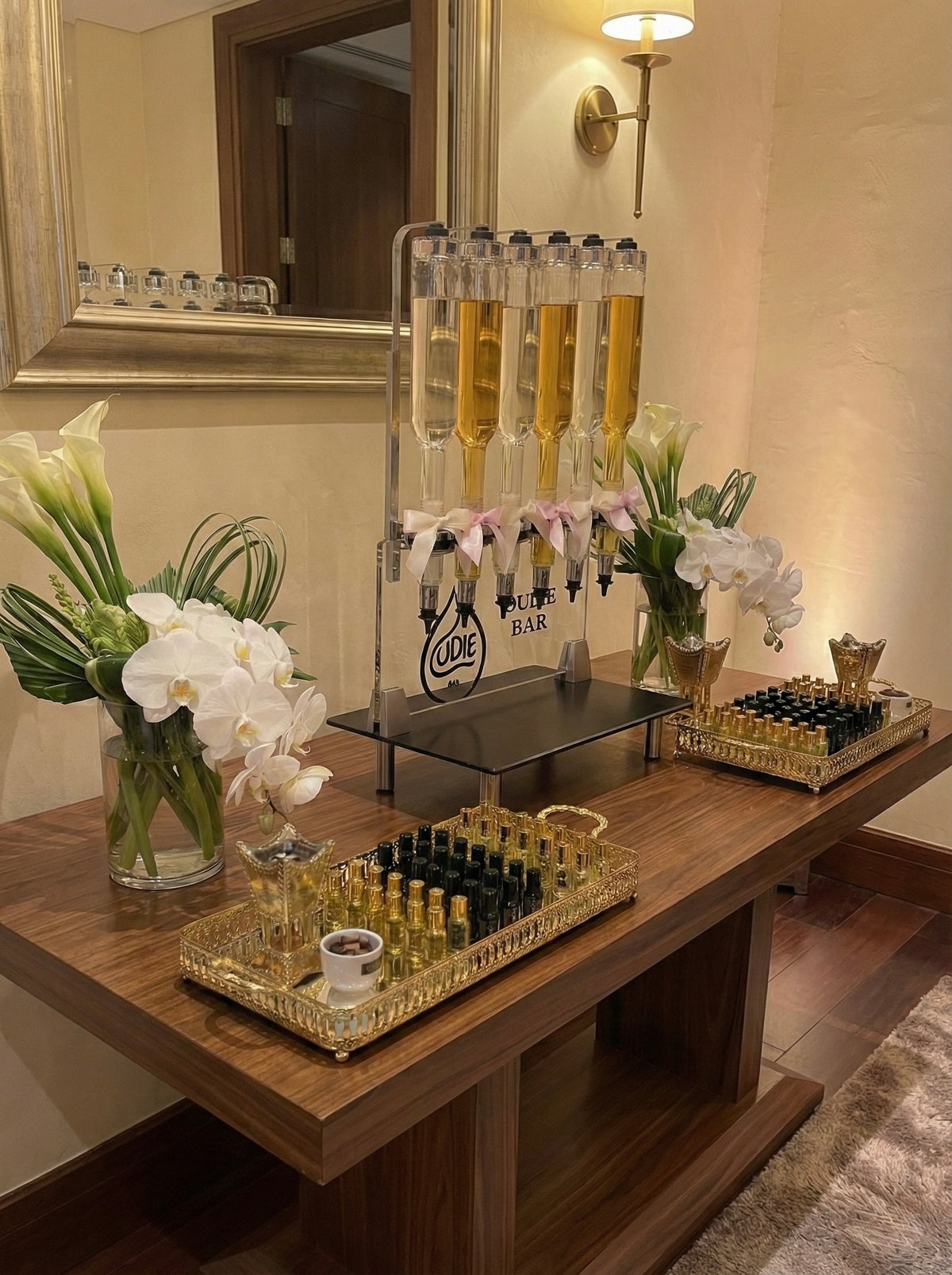A sophisticated Oud bar positioned elegantly against the wall of a high-end corporate venue. The setup features a sleek wooden console table holding the glass fragrance dispensers and gold serving trays, creating a refined side station that invites interaction without obstructing the room's flow. In the background, business professionals network in a modern conference atmosphere, highlighting the bar as a luxurious yet professional touch to the event.